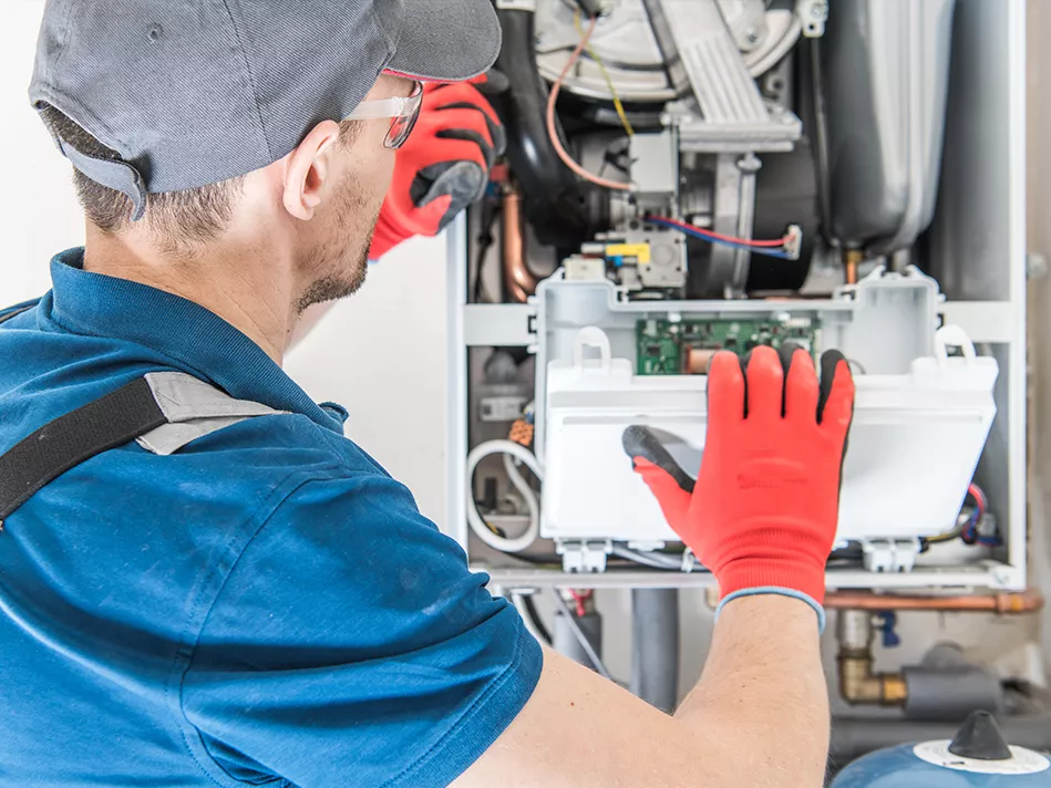 A white man in a blue short-sleeved polo, a grey baseball cap, red electrician gloves, and transparent safety goggles inspects hard wiring 
