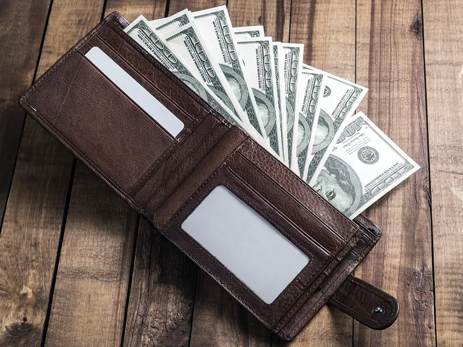 Brown leather wallet open and full of $100 bills rests on wood