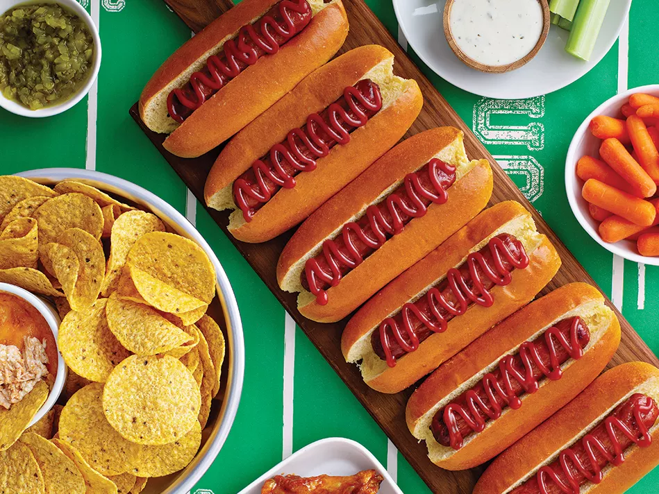 A diagonal row of hot dogs next to carrots, nachos, and other small dishes on a green and white tablecloth