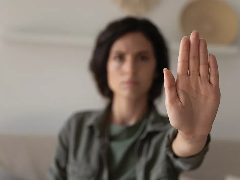A Hispanic woman with a dark brown bob and wearing a gray-green button-down shirt holds up her left hand, gesturing "stop"; only her left hand is in focus