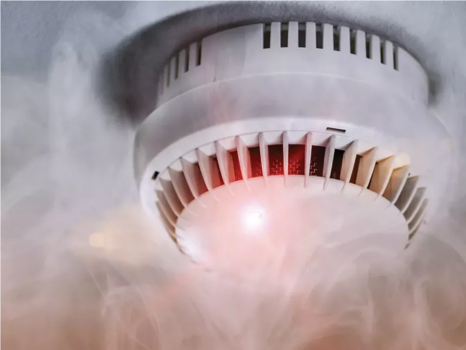 A white smoke alarm with a red light on and smoke around it