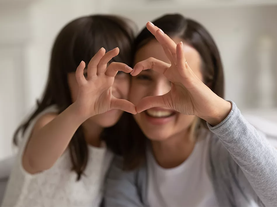 A white mom and daughter make a heart shape together, each one using one hand as half of the heart