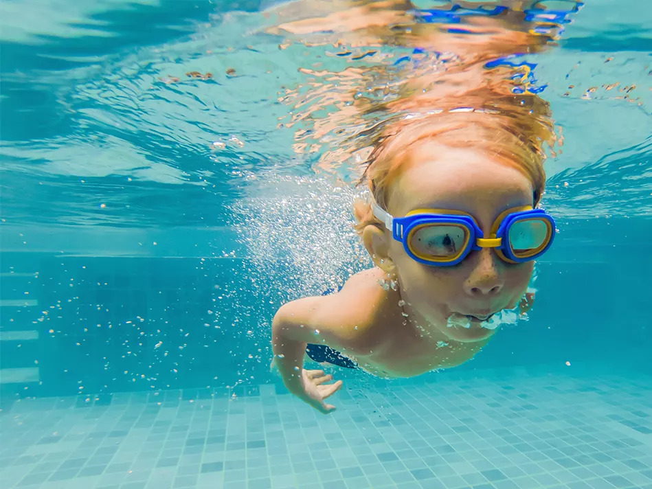 A young child in goggles swims underwater in a pool while blowing bubbles