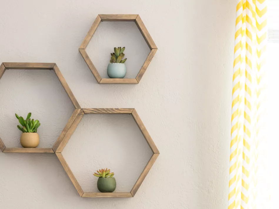 Honeycomb wall shelving made from light wood with a plant in the center of each of the 3 units adorn a beige wall to the left of a yellow and white curtain flanking light coming in through a window 