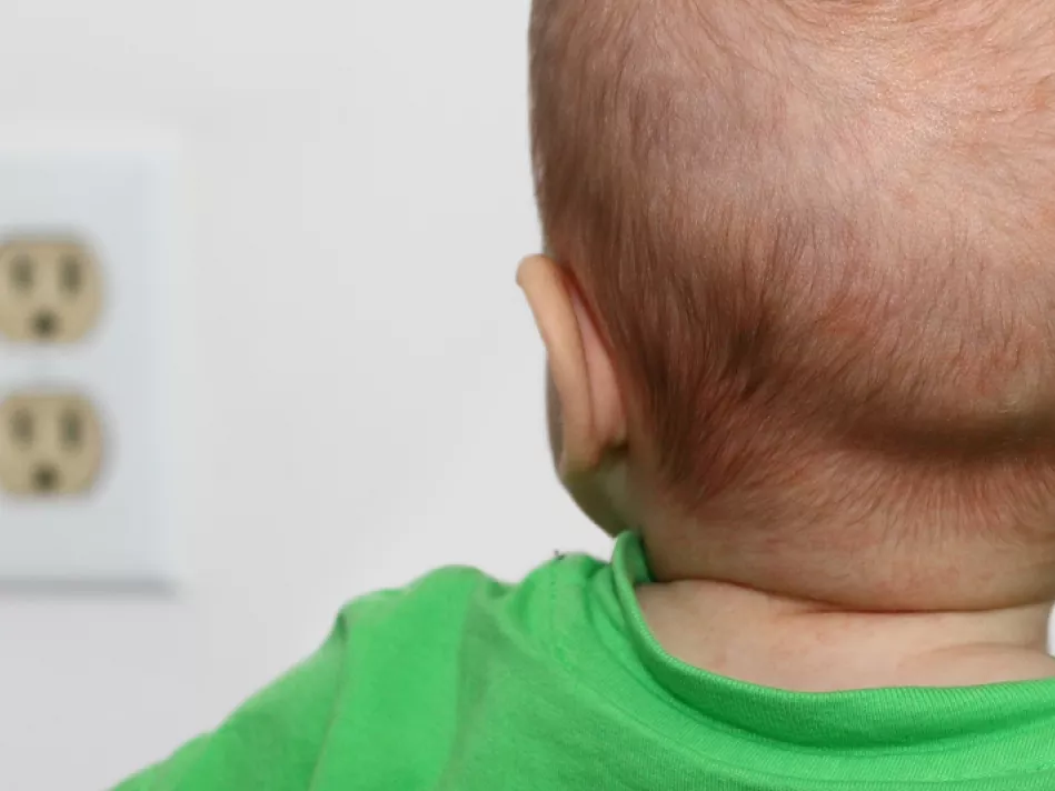 The back of the head of an infant in a green shirt who's staring at a power outlet