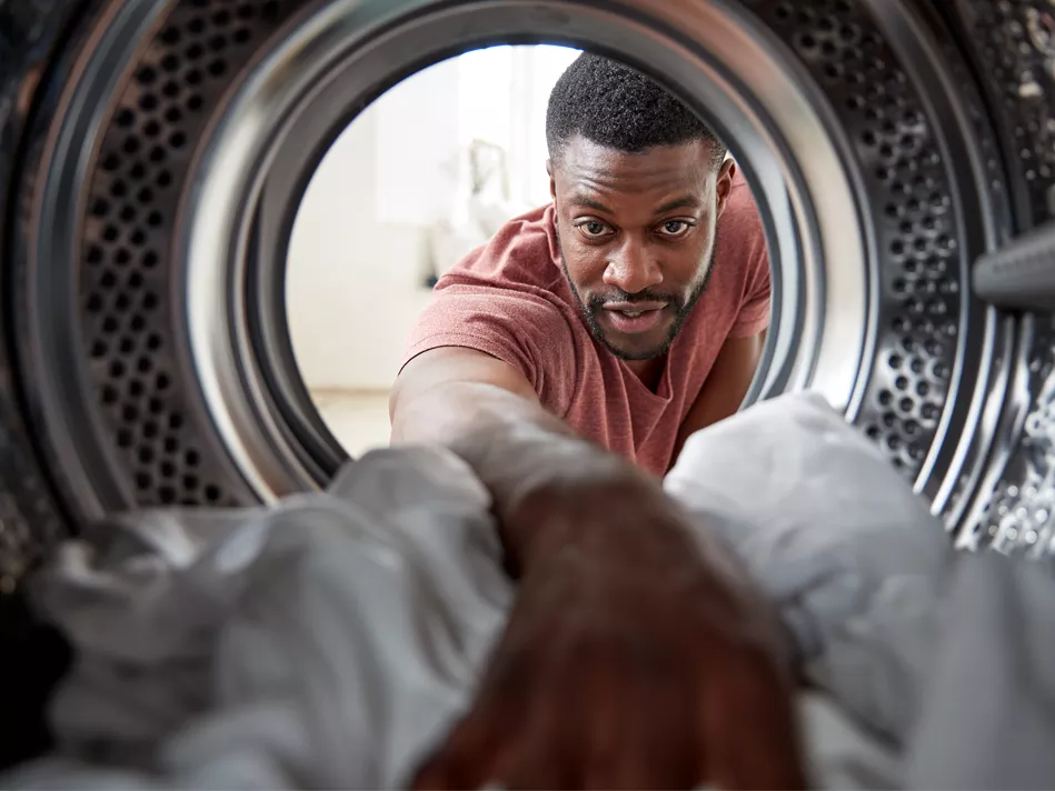 A Black man in a brownish-red shirt reaches into an open dryer with his right arm to grab a white garment