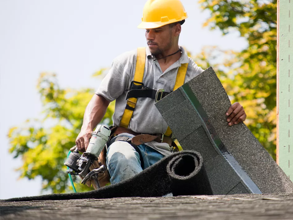 A black male in construction gear lays shingles on a roof