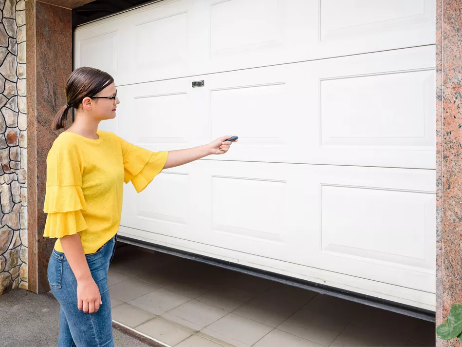 White woman in black glasses, yellow ruffled shirt, and jeans stands in front of white garage door while pressing garage door opener 