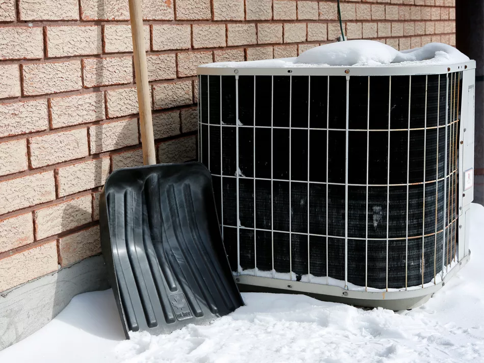 Snow piles up around an AC unit, while a shovel leans against the wall