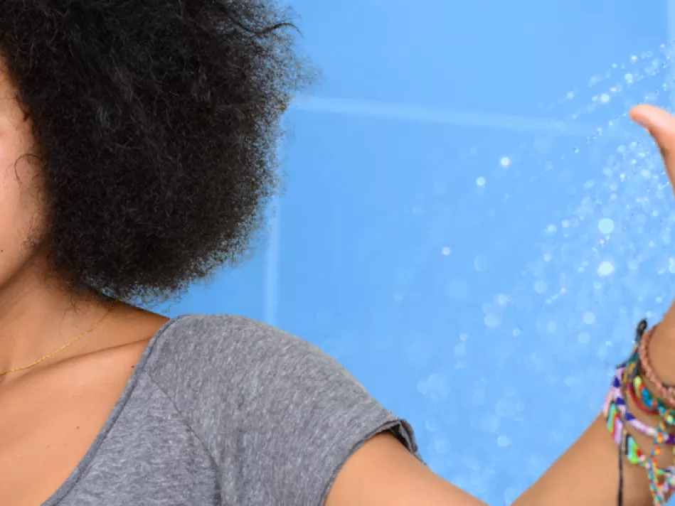 A Black woman in a gray shirt looks askance to the right with her left arm, which is adorned with colorful bracelets, is raised to the left; the background is a blue image of a showerhead with water coming out 