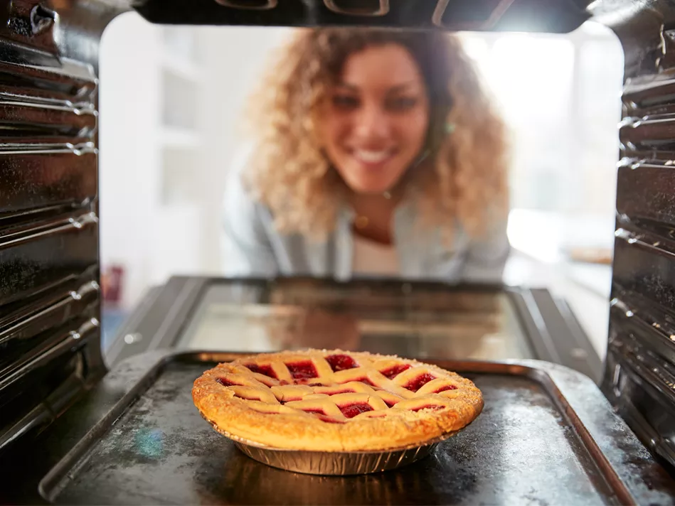 A Blended woman looks at a cherry pie sitting on an aluminum pan in a turned-on oven