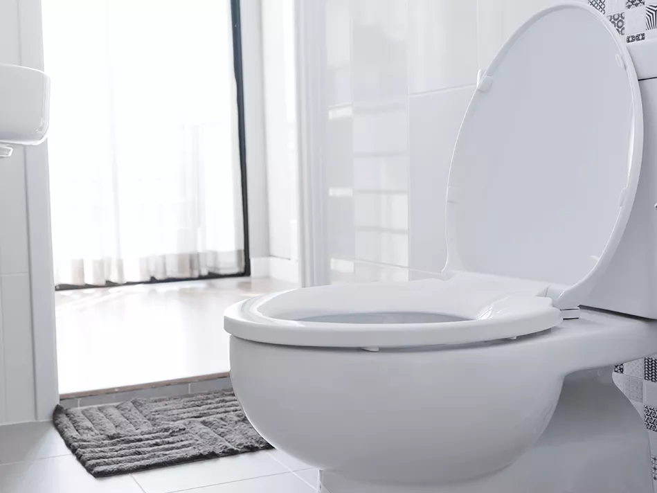 Light pours through a bedroom to show a white toilet in front of a gray mat and a white sink in a white-tiled en suite bathroom