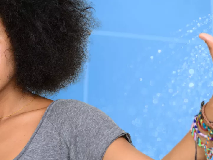 A Black woman in a gray shirt looks askance to the right with her left arm, which is adorned with colorful bracelets, is raised to the left; the background is a blue image of a showerhead with water coming out 