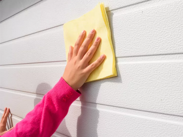 A light-skinned woman wearing a hot pink sweater with three buttons at the cuff of the sleeve wipes a white garage door with a folded light yellow cloth in her right hand