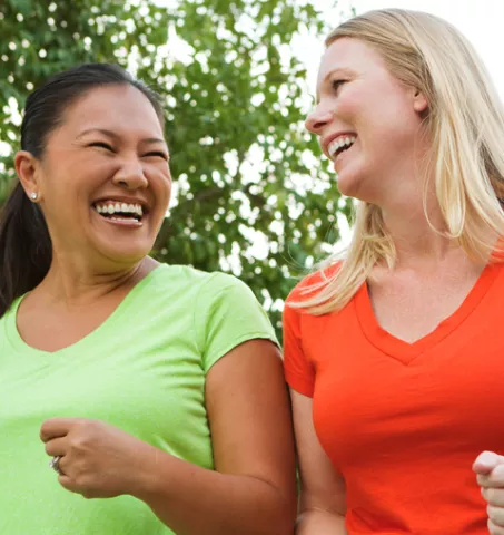 Una mujer asiática/isleña del Pacífico con una camisa verde lima riéndose con una mujer blanca con una camisa naranja rojiza