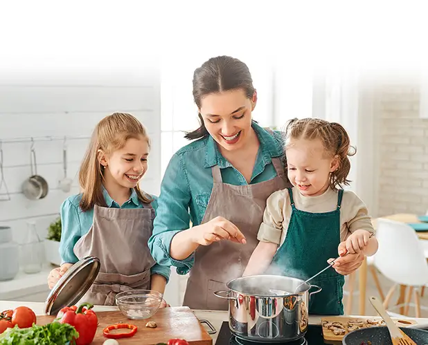 Young woman in orange shirt and gray pants kneeling beside a front-loading washing machine overflowing with soap suds, her hands are clasped to her face as she stares wide eyed in shock due to her broken washer machine, folded towels and detergent bottles on a wooden laundry cabinet, illustrating a big washing machine malfunction.